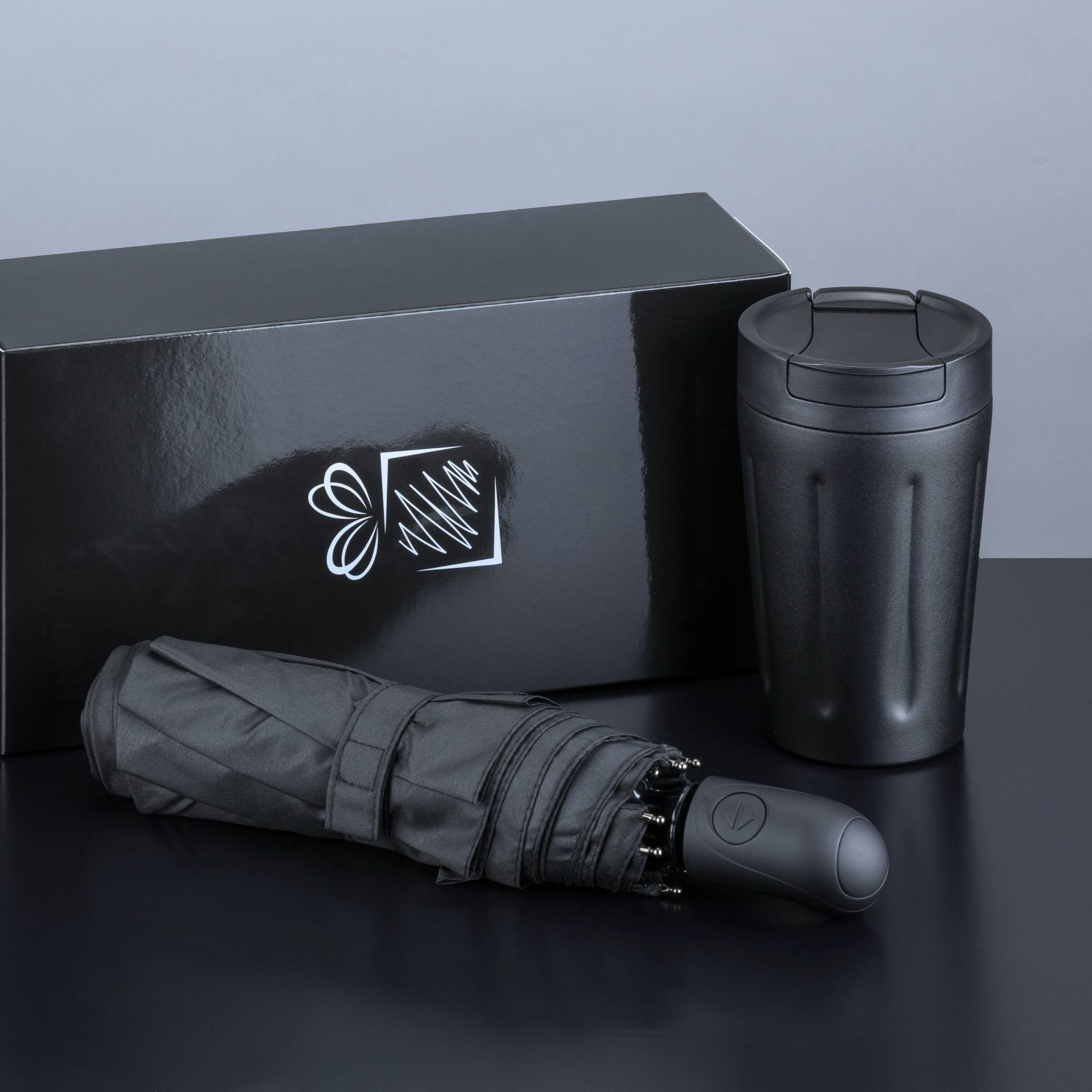 uoda dovanų dėžė su balta dovana simbolizuojančia emblema, šalia kurios guli sulankstomas juodas skėtis ir stovi juodas termo puodelis